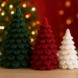 Puede incluir: Tres árboles de Navidad de ganchillo hechos a mano en verde, rojo y blanco. Los árboles están dispuestos sobre una superficie de madera con un fondo borroso de luces navideñas y un fondo rojo. Los árboles son de diferentes tamaños.