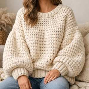 Könnte beinhalten: Cremefarbener, grob gestrickter Pullover mit rundem Ausschnitt und langen, voluminösen Ärmeln. Der Pullover hat ein strukturiertes, erhabenes Stichmuster und gerippte Bündchen. Das Model trägt blaue Jeans.