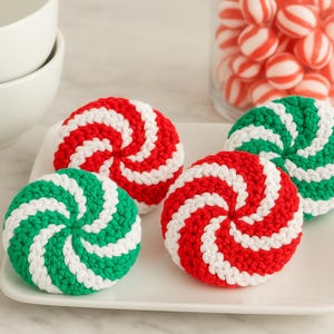 Puede incluir: Un conjunto de posavasos hechos a mano, tejidos a ganchillo, con un dise&ntilde;o de remolino de caramelo. Los posavasos son rojos, blancos y verdes, parecidos a caramelos de menta. Se muestran en un plato blanco.