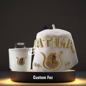 May include: A white custom fez hat and matching container. The hat features the name "FATIMA" in jeweled letters and an Egyptian-style emblem. The container has a handle and a similar emblem. The text "Custom Fez" is at the bottom.
