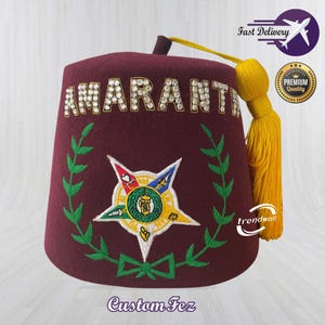 Puede incluir: Un fez burdeos con la palabra "AMARANT" en letras blancas adornadas. El sombrero presenta un emblema bordado con una estrella y hojas de laurel verdes. Una borla amarilla cuelga de un lado. El texto "Custom Fez" está en la parte inferior.
