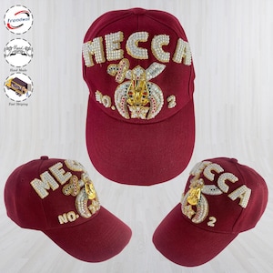 May include: A deep red baseball cap featuring the word MECCA in white pearl-like beads across the front. Below the text is a gold-toned emblem with colourful accents and the text NO. 2. The cap is shown from three different angles in the image.