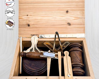 Op de afbeelding: Houten kist met diverse houtbewerkingsgereedschappen, waaronder een hamer, een passer en een liniaal. De gereedschappen zijn gemaakt van hout en messing, met een lichtblauw touwtje en een donkere houten basis. De kist is gemaakt van lichtgekleurd hout en heeft een messing sluiting.