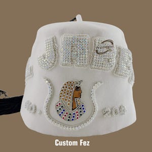 Peut inclure: Un fez blanc orné de broderies et de perles. Le chapeau présente un profil stylisé et le texte "Custom Fez" en bas. Il comprend un pompon noir et des éléments décoratifs. Le design comprend des accents perlés.
