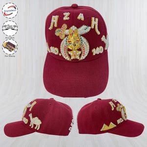 May include: A maroon baseball cap featuring an embroidered gold pharaoh bust, crossed swords, and pearl-like accents. The front displays the text AZAM, while the sides show a camel and pyramid motif, all crafted with intricate beadwork and embroidery.