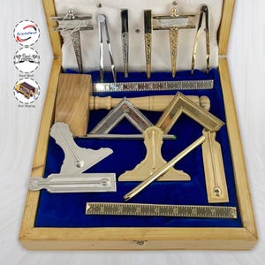 Puede incluir: Una caja de madera con interior de terciopelo azul contiene una colección de herramientas de dibujo antiguas. El juego incluye reglas, escuadras, compases y otros instrumentos en tonos plateados y dorados. La caja está abierta, revelando las herramientas bien dispuestas.