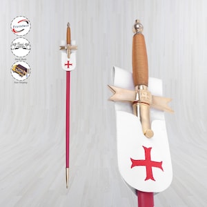 Puede incluir: Una espada decorativa con empuñadura de madera y guarda dorada en forma de cruz. La espada presenta un asta roja y un escudo blanco con un emblema de cruz roja. El diseño recuerda a las armas históricas.