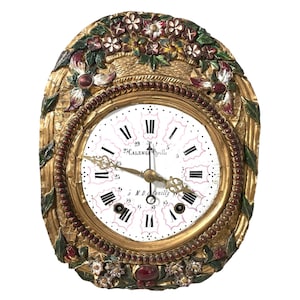 May include: A gold-toned oval clock with a white face and black Roman numeral hour markers. The clock has a floral design around the edge, with red, green, and white flowers. The clock face reads "Calenge Grilly" and "J.D. Montigny".