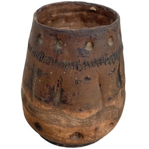 Puede incluir: Una olla de barro marrón con una boca ancha y un fondo redondeado. La olla tiene una superficie texturizada con líneas incisas y puntos.