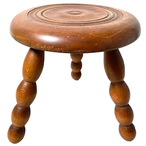 May include: A wooden stool with a round top and three legs. The legs are turned and have a series of rounded knobs.