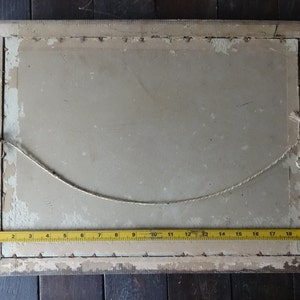 May include: The back of a framed picture with a white mat and a light brown wooden frame. A string is attached to the frame for hanging. A yellow measuring tape is placed along the bottom edge of the frame.