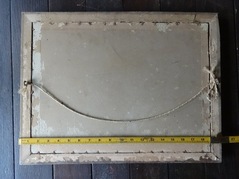 May include: The back of a framed picture with a white mat and a light brown wooden frame. A string is attached to the frame for hanging. A yellow measuring tape is placed along the bottom edge of the frame.