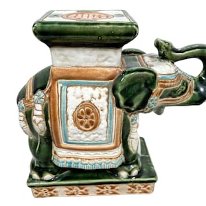 May include: A green ceramic elephant stool with white and brown accents. The elephant has a raised trunk and is decorated with intricate details. The stool has a square top and a base with a decorative pattern.