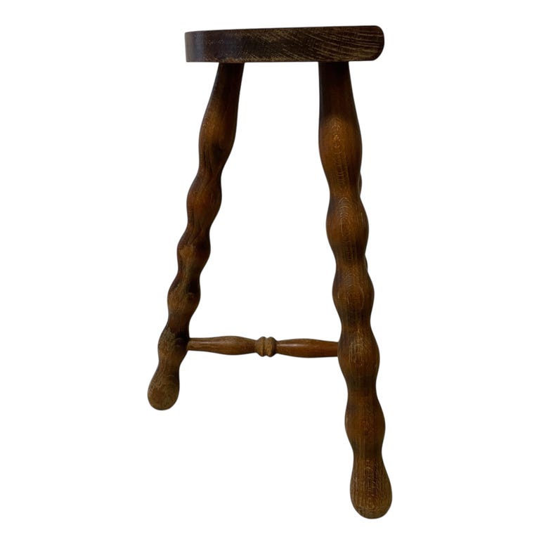 K&ouml;nnte beinhalten: Ein kleiner, antiker Holzschemel mit einer dunkelbraunen, oval geformten Sitzfl&auml;che. Die Beine sind aufwendig mit einem bauchigen Design geschnitzt und durch eine horizontale Strebe verbunden. Der Hocker ist in einem warmen, mittelbraunen Farbton gebeizt.