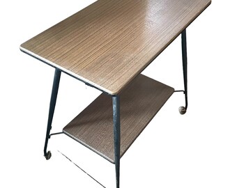 Puede incluir: Una mesa de madera de dos niveles con una tapa de laminado marrón y un estante inferior. La mesa tiene un marco de metal negro y cuatro ruedas.