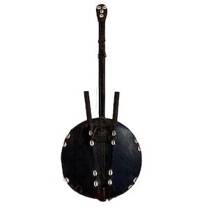 Può includere: Uno strumento a corde marrone scuro con un corpo rotondo e nero decorato con conchiglie bianche. Il manico lungo e sottile presenta un volto scolpito in cima. Due montanti verticali si estendono dal corpo.