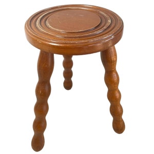 May include: A wooden stool with a round top and three legs. The legs are turned and have a rounded shape. The stool is made of light brown wood.