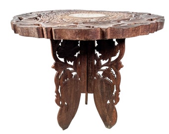 May include: A dark brown wooden folding table with intricate carvings in the shape of leaves and flowers. The table has a round top and three legs.