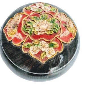 May include: A round wooden box with a floral design. The box is painted black with red, white, and green flowers. The flowers are carved into the wood and have a 3D effect.