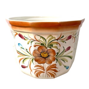 May include: A white ceramic planter with a brown rim and floral design. The planter features a large orange flower with yellow center and green leaves.