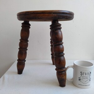 May include: A small, wooden stool with a round top and three turned legs. The stool is dark brown and has a rustic appearance.