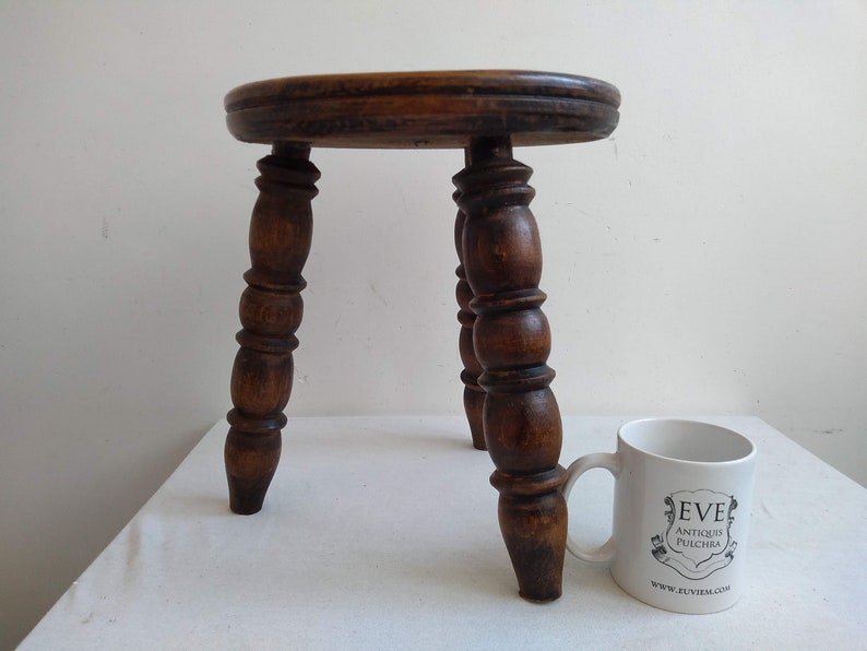 May include: A small, wooden stool with a round top and three turned legs. The stool is dark brown and has a rustic appearance.