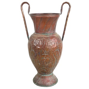 May include: A copper vase with two handles and an embossed floral design. The vase has a rustic, aged look.