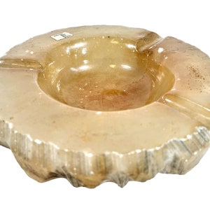May include: A white alabaster ashtray with a rough, textured exterior and a smooth, rounded interior. The ashtray has three indentations for cigarettes.