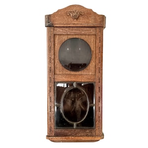 May include: A vintage wooden wall clock with a glass face and a carved wooden frame. The clock has a decorative design on the top and a small window on the bottom.