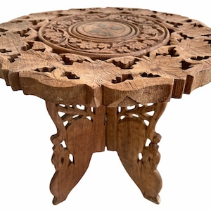 Könnte beinhalten: Ein kleiner, kunstvoll geschnitzter Holztisch mit runder Tischplatte und drei Beinen. Der Tisch ist aus hellbraunem Holz gefertigt und hat ein florales Design.