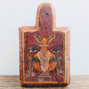 Może przedstawiać: Drewniana skrzynka z malowanym wzorem w kolorze czerwonym, pomarańczowym i żółtym. Wzór przedstawia stylizowaną twarz z dwoma oczami i motywem kwiatowym.