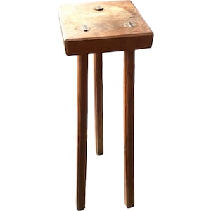 Może przedstawiać: Mały drewniany stołek z kwadratowym blatem i trzema nogami. Stołek jest wykonany z naturalnego drewna i ma rustykalny wygląd.