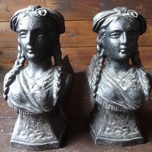 Peut inclure: Deux serre-livres en fonte de couleur gris foncé. Chaque serre-livre représente un buste de femme aux cheveux tressés et coiffée d'un bandeau. Les serre-livres ont une finition texturée et sont montés sur une base rectangulaire.