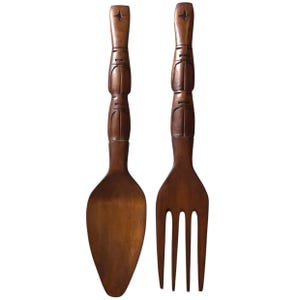 May include: A pair of wooden serving utensils, a spoon and a fork, with carved details. The handles are intricately designed with a starburst at the top. The utensils are a warm brown color, suggesting a natural wood finish.