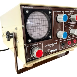 Puede incluir: Un osciloscopio beige y marrón con una pantalla que muestra un patrón de cuadrícula. El dispositivo tiene perillas para ajustar configuraciones como "Cadrage", "Balayage" y "Gain". También tiene una etiqueta que dice "Oscillographe PC 774" y "Jeulin".