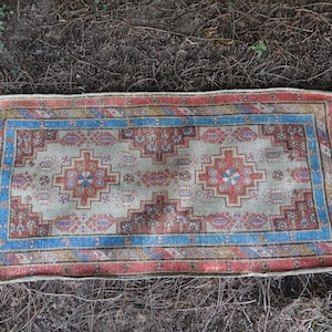 May include: A vintage Turkish rug with a cream background and a colorful geometric pattern in shades of red, blue, and orange. The rug has a fringed edge and is lying on a bed of brown pine needles.