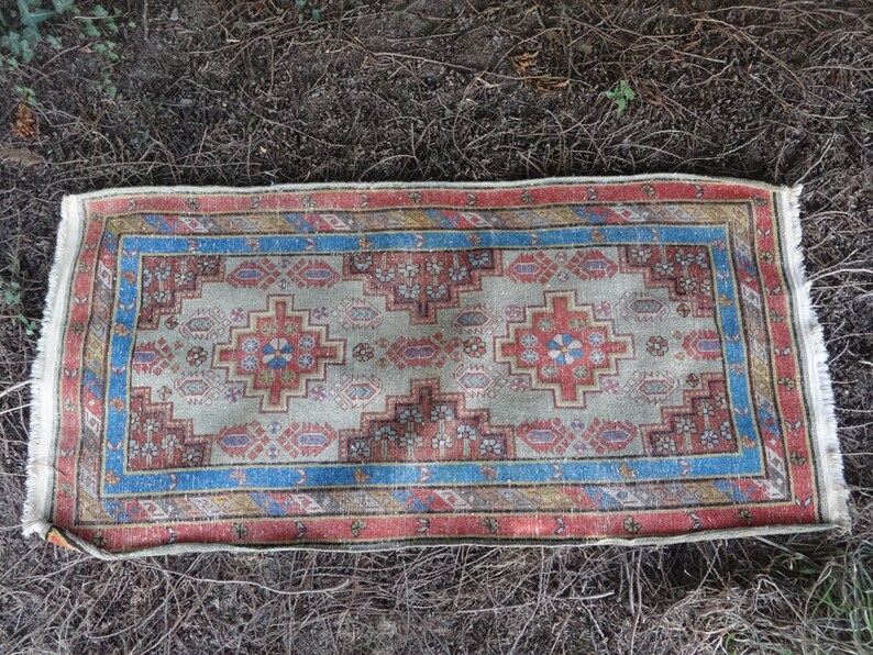 May include: A vintage Turkish rug with a cream background and a colorful geometric pattern in shades of red, blue, and orange. The rug has a fringed edge and is lying on a bed of brown pine needles.