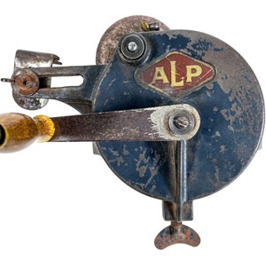 Könnte beinhalten: Vintage handbetriebenes Schleifwerkzeug mit dunkelblauem, kreisförmigem Gehäuse und Holzgriff. Das Werkzeug verfügt über eine Schleifscheibe und einen Metallarm. Die Buchstaben "ALP" sind auf der Vorderseite sichtbar. Zum Schärfen von Klingen.