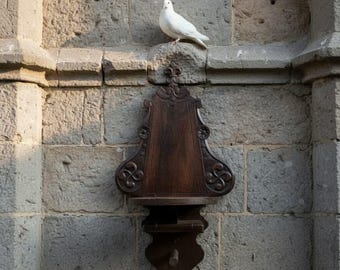 May include: A white dove perched on a stone ledge above a dark wooden wall-mounted decorative piece with ornate carvings. The piece is attached to a gray stone brick wall.