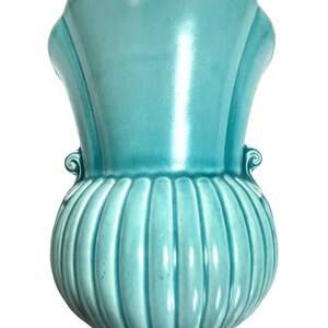 May include: A turquoise ceramic vase with a fluted, bulbous base and a flared top. The vase has two small, decorative handles and a glossy finish. The overall shape is reminiscent of a stylized flower or a classic urn.