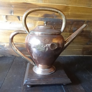 Puede incluir: Una tetera de cobre con asa y pico de latón, sentada sobre una base de madera. La tetera tiene un cuerpo redondeado y un pico ligeramente acampanado.