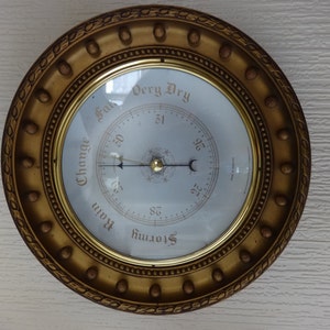 May include: A gold-framed barometer with a white face and black hands. The face has a scale from 28 to 31, with markings for "Stormy", "Rain", "Change", "Fair", and "Very Dry". The barometer is made in England.