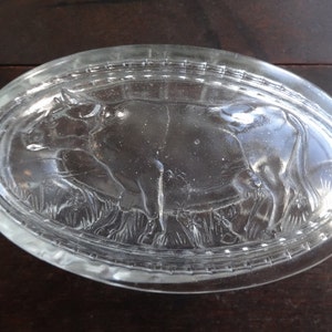May include: A clear glass butter dish with a raised design of a cow in a field. The dish is oval shaped and has a textured rim.