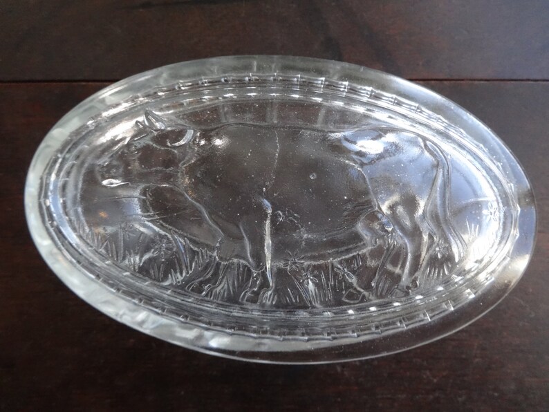 May include: A clear glass butter dish with a raised design of a cow in a field. The dish is oval shaped and has a textured rim.