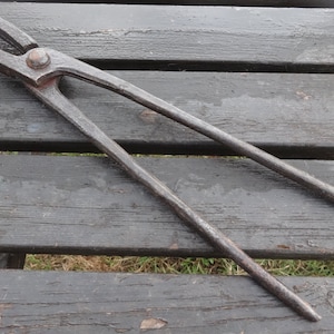 Puede incluir: Un par de tenazas de herrero de metal negro con una cabeza redonda. Las tenazas están sobre una superficie de madera.