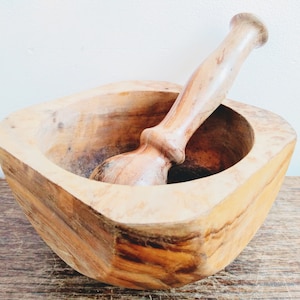 May include: A wooden mortar and pestle set. The mortar is square shaped and made of light brown wood. The pestle is made of the same wood and has a rounded end.