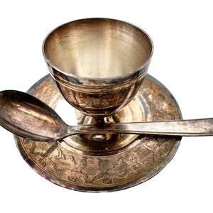 May include: A silver egg cup and spoon set. The egg cup has a round, bowl-shaped top and a pedestal base. The spoon is small and has a rounded bowl.