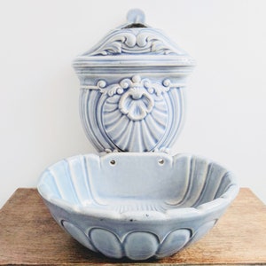 May include: A light blue ceramic wall planter with a decorative top and a scalloped bottom. The planter has two holes for hanging.