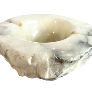Op de afbeelding: Een witte albasten asbak met een ruw, getextureerd exterieur. De asbak heeft een ronde opening in het midden en een licht verhoogde rand.