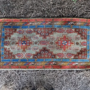 May include: A traditional rug with a geometric pattern in shades of red, blue, and beige. The rug has a white fringe border.